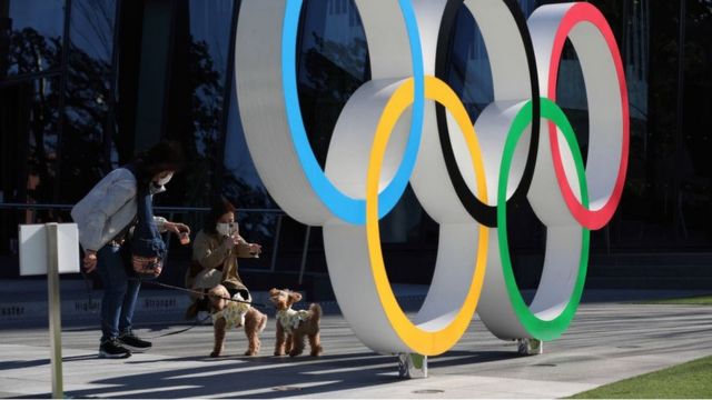 People interact on a public bench behind the Olympic symbol