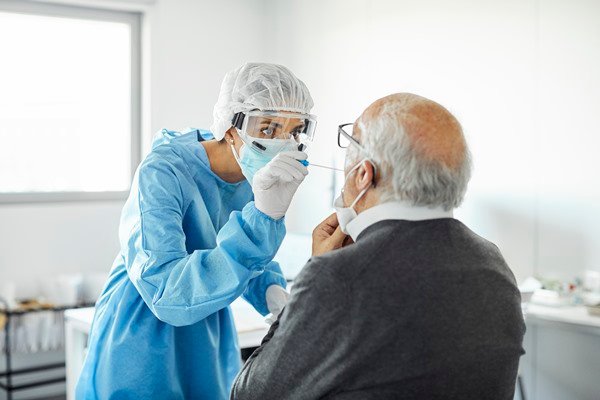 In the color photo, a person wearing blue places a cotton swab in the mouth of a seated elderly man.