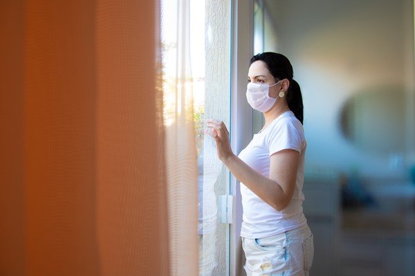 In the color photo, a woman looks out the window.  She is standing and wearing a mask