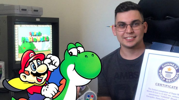 Imagem: Foto de Matheus Furtado segurando o certificado do Guinness numa mão e controle de SNEA na outra, com uma TV ligada no Super Mario World ao fundo e na frente uma imagem promocional de Mario montado no Yoshi.